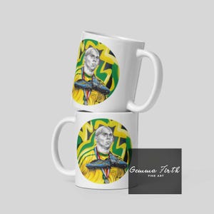 Puede incluir: Dos tazas de cer&aacute;mica blanca apiladas una encima de la otra. Las tazas presentan una ilustraci&oacute;n colorida de un jugador de f&uacute;tbol sosteniendo un par de botas de f&uacute;tbol. El texto "Gemma Firth Fine Art" est&aacute; impreso en la taza de abajo.