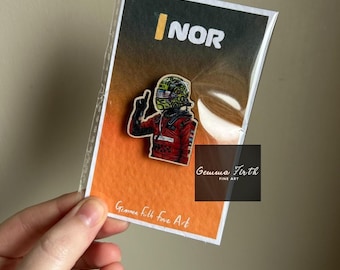 Puede incluir: Un pin con un piloto de Fórmula 1 con traje de carreras rojo, casco y guantes, haciendo un gesto con la mano. El pin está en una tarjeta con el texto "NOR" y "Gemma Firth Fine Art". El fondo es naranja y negro.
