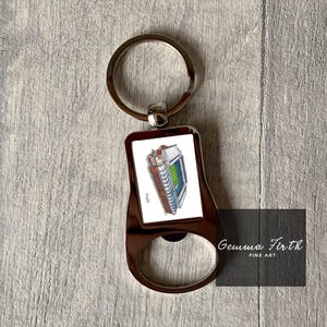 May include: A silver bottle opener keychain with a white rectangular insert featuring a color illustration of a stadium. The keychain has a silver ring and the text "Gemma Firth Fine Art" is printed on the bottom.
