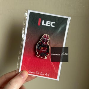 May include: A pin badge depicting a Formula 1 driver in a red racing suit and helmet. The pin is on a card with the text "LEC" and "Gemma Firth Fine Art". The pin is in a clear plastic sleeve.
