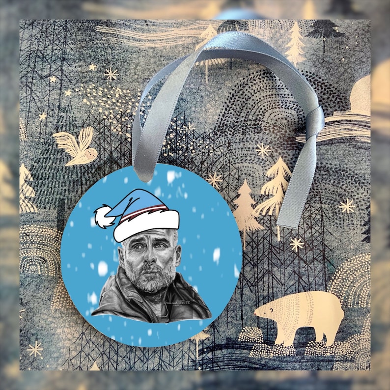 May include: Circular Christmas tree decoration with a portrait of a man in a Santa hat, set against a light blue backdrop with falling snowflakes. It is attached to a light blue ribbon. The background has a winter forest design.