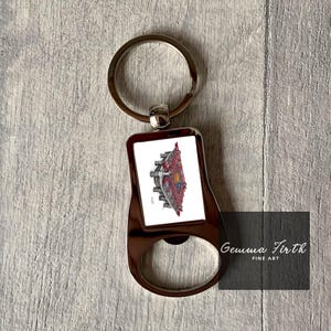 May include: A silver-colored bottle opener keychain with a rectangular image of a red and gold building. The keychain has a ring for keys and a bottle opener on the bottom. The image has the text "Gemma Firth FINE ART".
