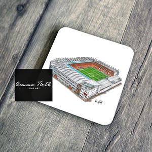 May include: A square coaster featuring a detailed illustration of a stadium with a green field and orange seating. The coaster has the text "G.F.C." and is set against a light wood grain background.