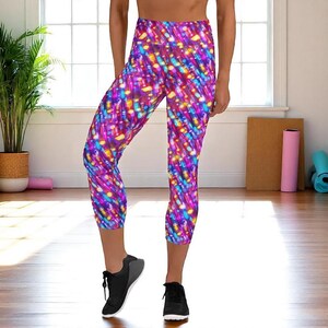 Może przedstawiać: Żywe, wielokolorowe legginsy capri z dynamicznym, abstrakcyjnym wzorem. Legginsy mają wysoki stan i skróconą długość, idealne do ćwiczeń lub noszenia na co dzień. Projekt zawiera odcienie różu, fioletu, niebieskiego i żółtego, tworząc efekt wizualny. Legginsy są połączone z czarnymi butami sportowymi.
