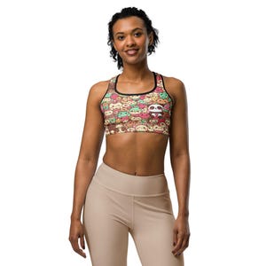 May include: A colorful sports bra featuring a pattern of cartoon characters in various colors. The bra has a black trim and straps. The design includes a panda bear, strawberries, and other cute illustrations. The model is wearing beige leggings.