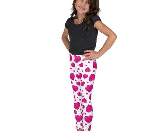 May include: White leggings patterned with pink hearts. The leggings are worn by a person wearing a black t-shirt. The hearts vary in size and are scattered across the leggings. The person is smiling with their hands on their hips.