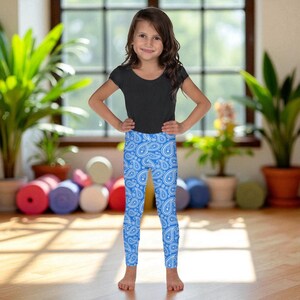 Peut inclure: Une enfant porte un haut noir et des leggings bleus à motifs cachemire blancs. Les leggings sont moulants et arrivent aux chevilles. L'arrière-plan comprend des plantes et une fenêtre.