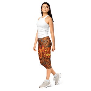 May include: A white sleeveless ribbed tank top paired with orange and brown leggings featuring a repeating pattern of jack-o'-lanterns, flames, bats, and swirling decorative elements. The leggings extend to mid-calf length and are worn with grey sneakers.