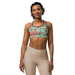 May include: A sports bra with a colorful floral and geometric pattern in red, green, black, and white. The bra has a black trim and straps. The model is wearing beige leggings.
