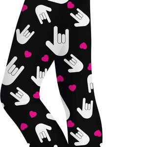 Puede incluir: Leggings negros con un patrón repetido de gestos de manos 'rock on' blancos y corazones rosas. Los leggings son ajustados y adecuados para diversas actividades.