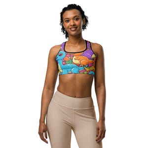 May include: A colorful sports bra featuring a cartoon dinosaur design. The bra has a black trim and straps, with a vibrant pattern of blue, orange, and purple dinosaurs on a multicolored background. The model is wearing beige leggings.