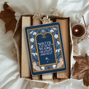Puede incluir: Una tarjeta rectangular azul con el texto "Winter is the time for reading, creativity, and rest" se encuentra sobre una pila de libros. La tarjeta presenta un diseño floral con dos zorros. Una vela encendida y hojas de otoño están en el fondo.