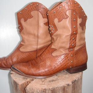 May include: A pair of brown cowboy boots with a textured, crocodile-like pattern. The boots have a light brown upper section and are adorned with silver studs. The toe of each boot is reinforced with a silver metal cap.