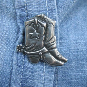 May include: A silver cowboy boot pin with a star on the front. The pin is attached to a blue denim shirt.