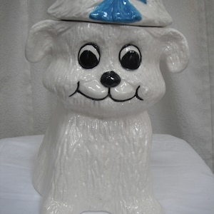 May include: White ceramic cookie jar shaped like a dog wearing a blue bow. The dog has large black eyes and a black nose.