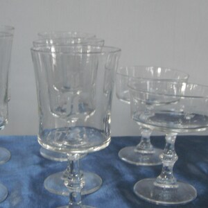 May include: A collection of clear glass stemware, including tall water goblets, short tumblers, and coupe glasses. The glasses have a classic design with a rounded base and a slender stem. The glassware is arranged on a blue surface.