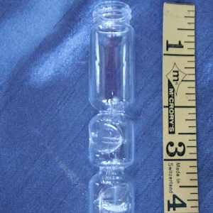 May include: A clear glass vial with a unique, segmented design. The vial is approximately 4 inches tall, with a series of rounded sections connected by narrower necks. A yellow and black measuring tape is next to the vial.