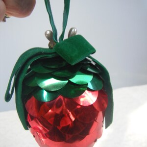 May include: A festive Christmas ornament shaped like a strawberry, featuring red and green sequins. The ornament has a green velvet top and a green ribbon for hanging. Two small pearl-like beads are attached to the top.