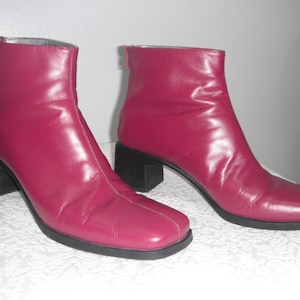 May include: A pair of burgundy ankle boots with a square toe and a black block heel. The boots have a zipper on the back and are made of a smooth, shiny material. The heel height is approximately 2 inches.