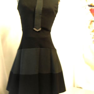May include: A sleeveless black dress with a high collar and a decorative strip down the front. The dress features a contrasting panel design on the skirt, creating a layered effect. The dress is displayed on a mannequin.