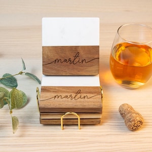 May include: A set of personalized coasters with a white and wood grain design, featuring the name "martin" in a cursive font. A glass of amber liquid, a cork, and a gold-toned coaster holder are also present.