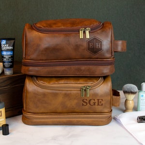 May include: Two brown leather travel toiletry bags with zipper closures and a top handle. The top bag has the initials "E.H." embossed on the front. The bottom bag has the initials "S.G.E." embossed on the front.