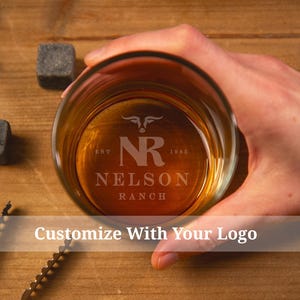 May include: A rocks glass filled with amber liquid, featuring the etched text "NR NELSON RANCH" and a bull head logo. Two square whiskey stones and a bar tool are on a wooden surface. Text at the bottom reads "Customize With Your Logo."