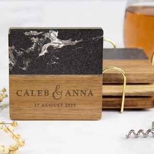 May include: Square wooden coasters with a black and white marble design on top and engraved with "CALEB & ANNA" and "17 AUGUST 2025". A set of coasters is in a gold-colored holder. A glass of liquid and a corkscrew are in the background.