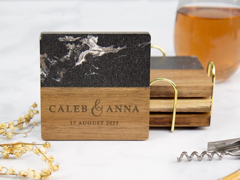 May include: Square wooden coasters with a black and white marble design on top and engraved with "CALEB & ANNA" and "17 AUGUST 2025". A set of coasters is in a gold-colored holder. A glass of liquid and a corkscrew are in the background.