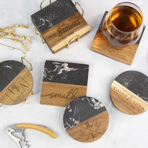 May include: A collection of personalized coasters with a black and white marble design paired with wooden accents. Some coasters feature engraved names and dates, while others have initials. A wine glass, cork, and corkscrew are also present.