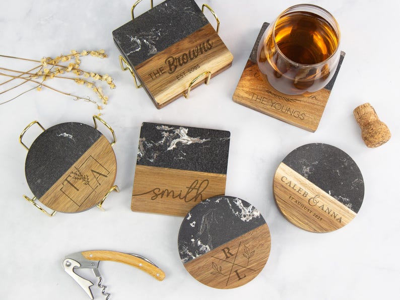 May include: A collection of personalized coasters with a black and white marble design paired with wooden accents. Some coasters feature engraved names and dates, while others have initials. A wine glass, cork, and corkscrew are also present.
