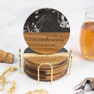 May include: A set of personalized coasters with a black and white marbled top and wooden base. The coasters are engraved with the names "Caleb & Anna" and the date "17 August 2025". They are displayed in a gold metal holder.