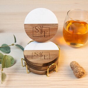 May include: A set of four round coasters with a white and wood grain design, stacked in a gold metal holder. Each coaster has a floral design and the letters "S" and "E" inside a square. A glass of amber liquid and a cork are also present.