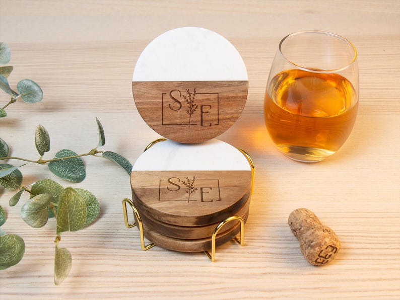 May include: A set of four round coasters with a white and wood grain design, stacked in a gold metal holder. Each coaster has a floral design and the letters "S" and "E" inside a square. A glass of amber liquid and a cork are also present.
