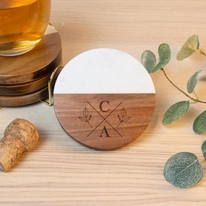 May include: A set of wooden coasters with a white marble top half and a brown wooden bottom half. The bottom half is engraved with the letters "C" and "A" and floral accents. A glass of liquid sits on top of a coaster. A cork and gold coaster holder are also present.