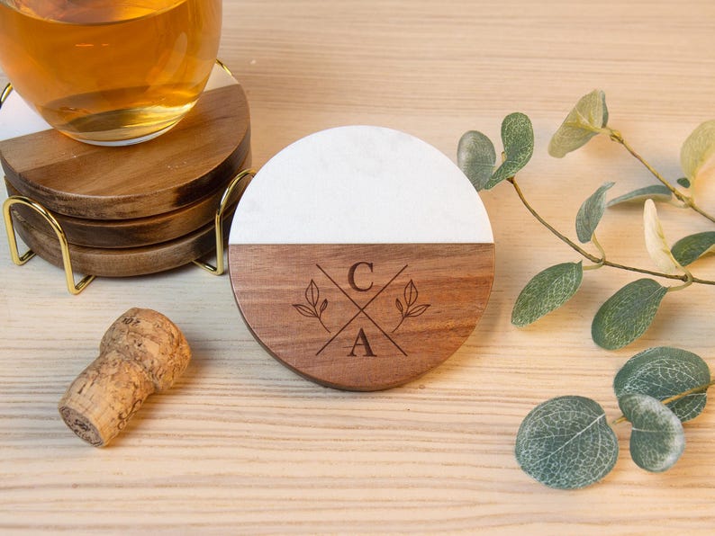 May include: A set of wooden coasters with a white marble top half and a brown wooden bottom half. The bottom half is engraved with the letters "C" and "A" and floral accents. A glass of liquid sits on top of a coaster. A cork and gold coaster holder are also present.