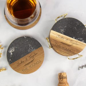 May include: Round coasters with a two-tone design, featuring a section of dark gray marble and a section of light wood. The coasters are engraved with the names "Jake & Taylor" and "THE YOUNGS". One coaster has a gold metal handle.