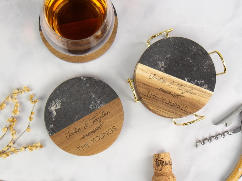 May include: Round coasters with a two-tone design, featuring a section of dark gray marble and a section of light wood. The coasters are engraved with the names "Jake & Taylor" and "THE YOUNGS". One coaster has a gold metal handle.