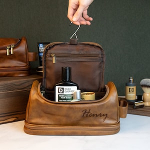 May include: A brown leather toiletry bag with the name "Henry" engraved on the front. The bag is open, revealing a black bottle of "Duke Cannon" hair wash and other grooming products. A hand holds the bag's hanging hook.