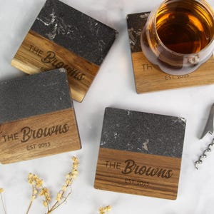 May include: Square coasters with a two-tone design. The top half is dark gray stone, and the bottom half is wood. The wood portion is engraved with "The Browns" and "EST. 2025". A glass of amber liquid sits on one coaster.