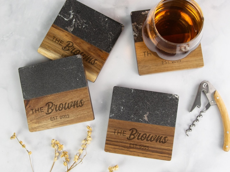 May include: Square coasters with a two-tone design. The top half is dark gray stone, and the bottom half is wood. The wood portion is engraved with "The Browns" and "EST. 2025". A glass of amber liquid sits on one coaster.