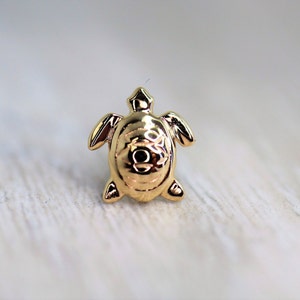 May include: A small, gold-colored turtle figurine. The turtle has a detailed shell design and is posed with its flippers extended. The figurine is set against a light-colored, textured background.