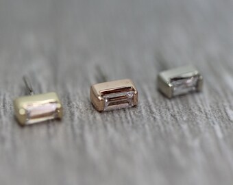 May include: Three small, rectangular-shaped earrings with a clear, faceted stone set in the centre. The earrings are shown in three different metal finishes: gold, rose gold, and silver.