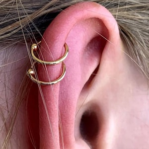 May include: Two gold hoop earrings with small ball accents. The earrings are worn in the upper ear cartilage.
