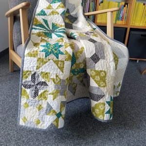 May include: A patchwork quilt with a variety of geometric patterns in shades of green, gray, and white. The quilt is folded and draped over a wooden armchair.