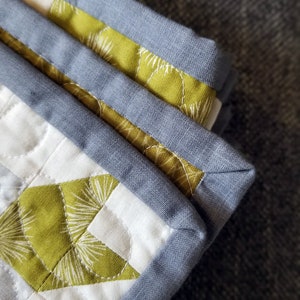 May include: Close-up of a quilt with a geometric pattern in shades of green, white, and gray. The quilt is edged with a gray fabric binding.