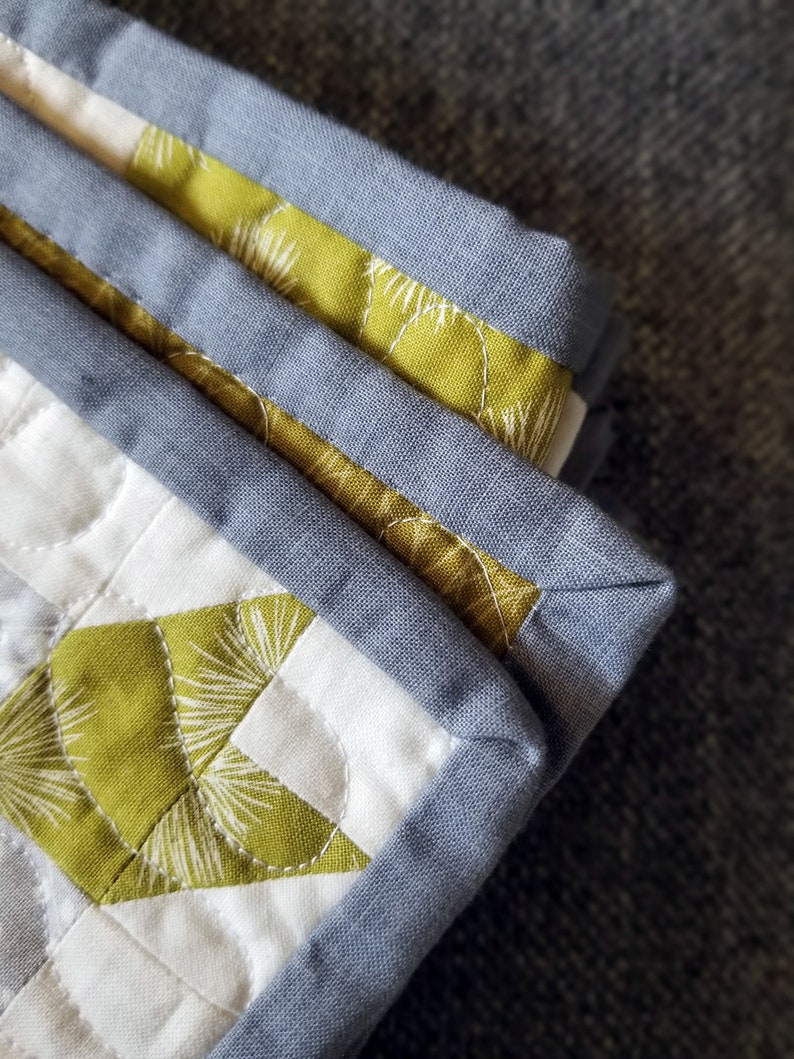 May include: Close-up of a quilt with a geometric pattern in shades of green, white, and gray. The quilt is edged with a gray fabric binding.