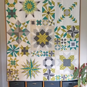 May include: A white quilt with a patchwork design featuring various geometric shapes in shades of green, gray, and white. The quilt is hanging on a wall with wooden shelves and storage bins below.