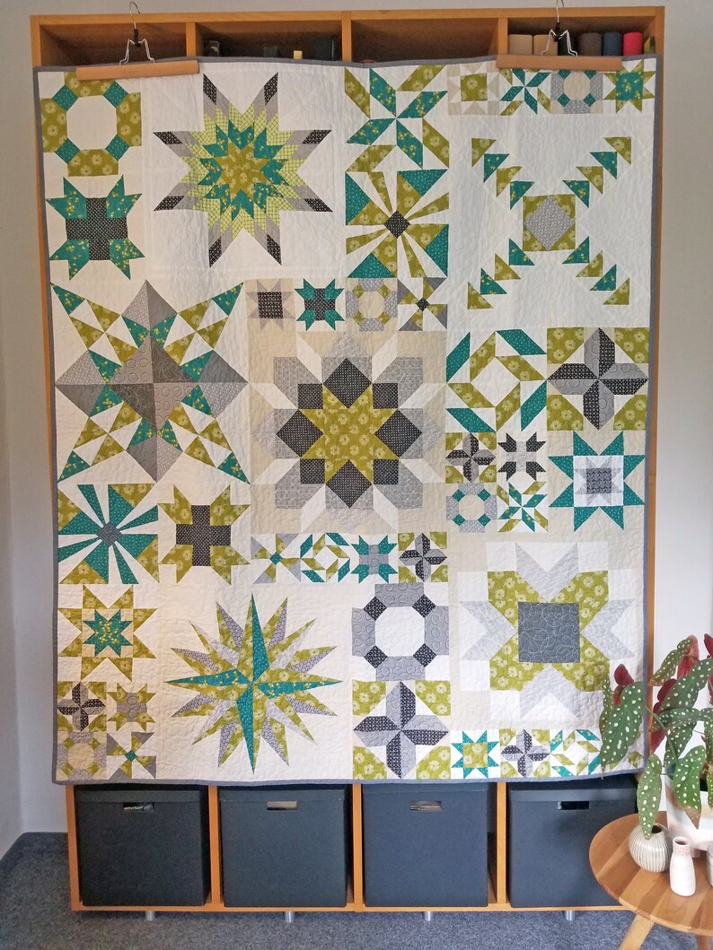 May include: A white quilt with a patchwork design featuring various geometric shapes in shades of green, gray, and white. The quilt is hanging on a wall with wooden shelves and storage bins below.