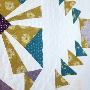 May include: A quilt with a geometric pattern in shades of teal, gold, purple, and white. The quilt is made of fabric with a variety of patterns, including polka dots, floral designs, and geometric shapes.